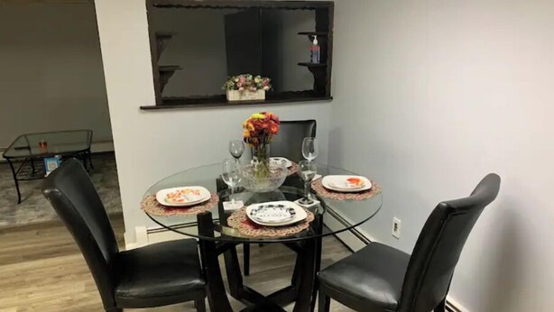 Un coin repas avec une table en verre entourée de quatre chaises noires, un vase avec des fleurs et des assiettes avec des couverts.