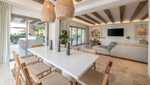 A modern living room with a white dining table, wooden chairs, and a sofa. There are two large woven pendant lights, a flat-screen TV mounted on the wall, and a large window offering a view of the outdoors. - Luxury 3 Bedroom Beachfront Cancun Vila  (Punta Sam)