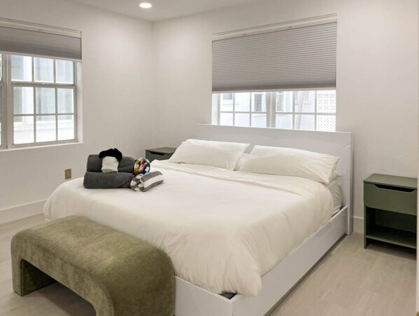 A modern bedroom with a large bed, two bedside tables, a bench, and a window with blinds. - Marley SoFi: Walk to beach, 3 Beds, Heated POOL (Miami Beach)