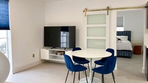 Een moderne hotelkamer met een witte eettafel, blauwe stoelen, een flatscreen-tv, een bed met een blauwe hoofdeinde en een schuifdeurkast.