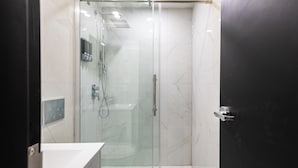 Un baño moderno con ducha de vidrio, lavamanos blanco y puerta oscura.