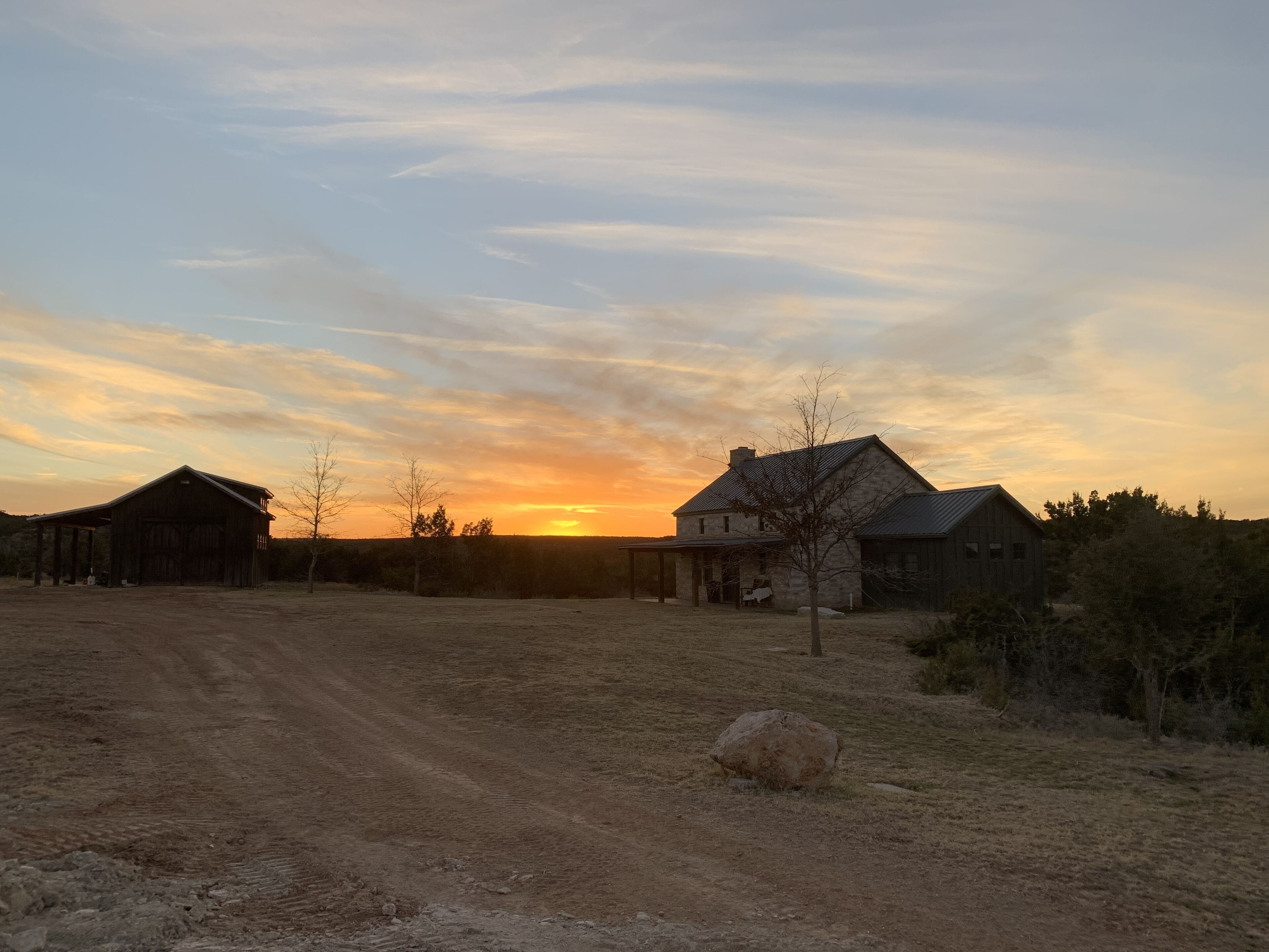 A rural landscape with a dirt road, a barn, and a house during sunset.