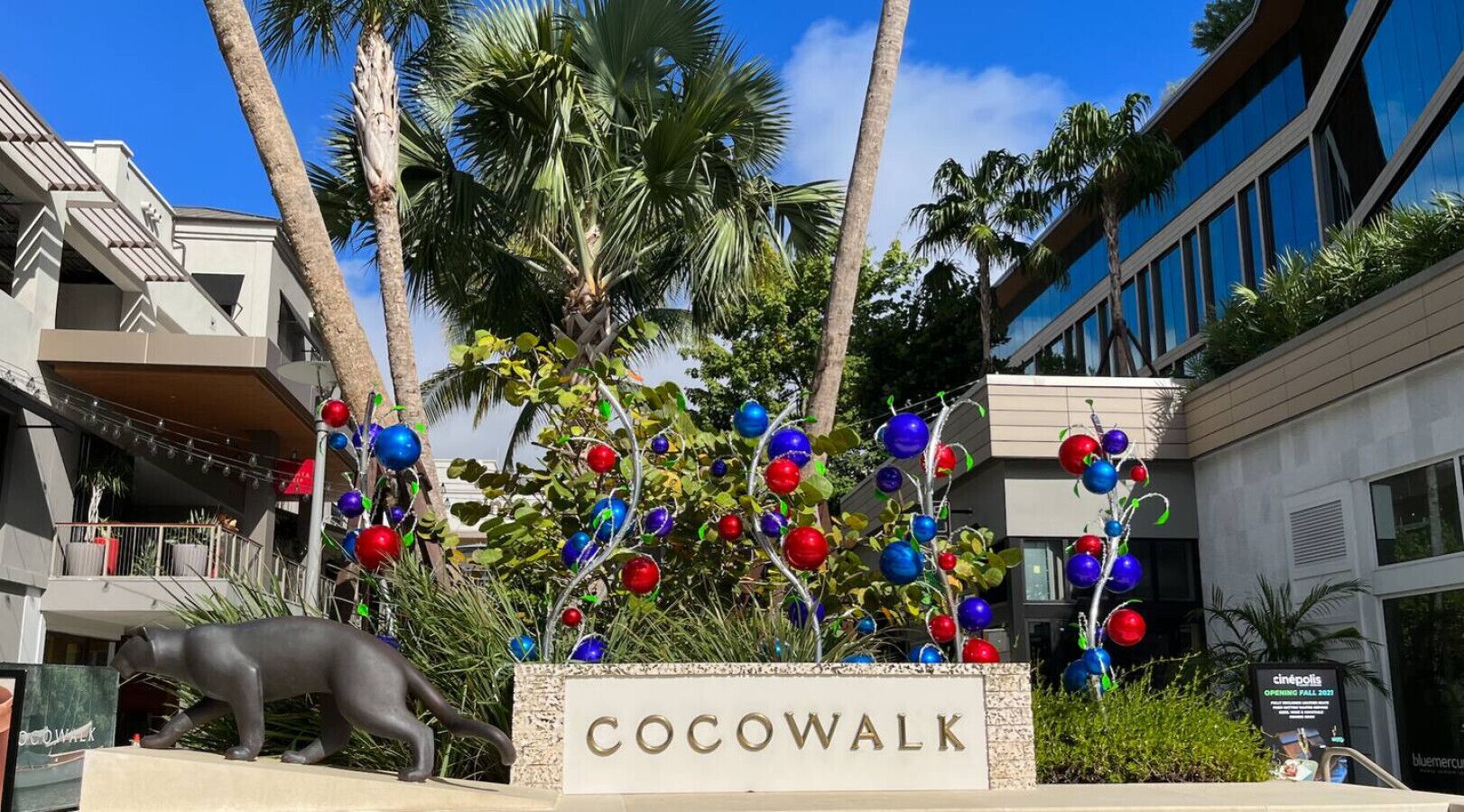 Señal para 'Cocowalk' con un árbol decorativo y una escultura de un perro.