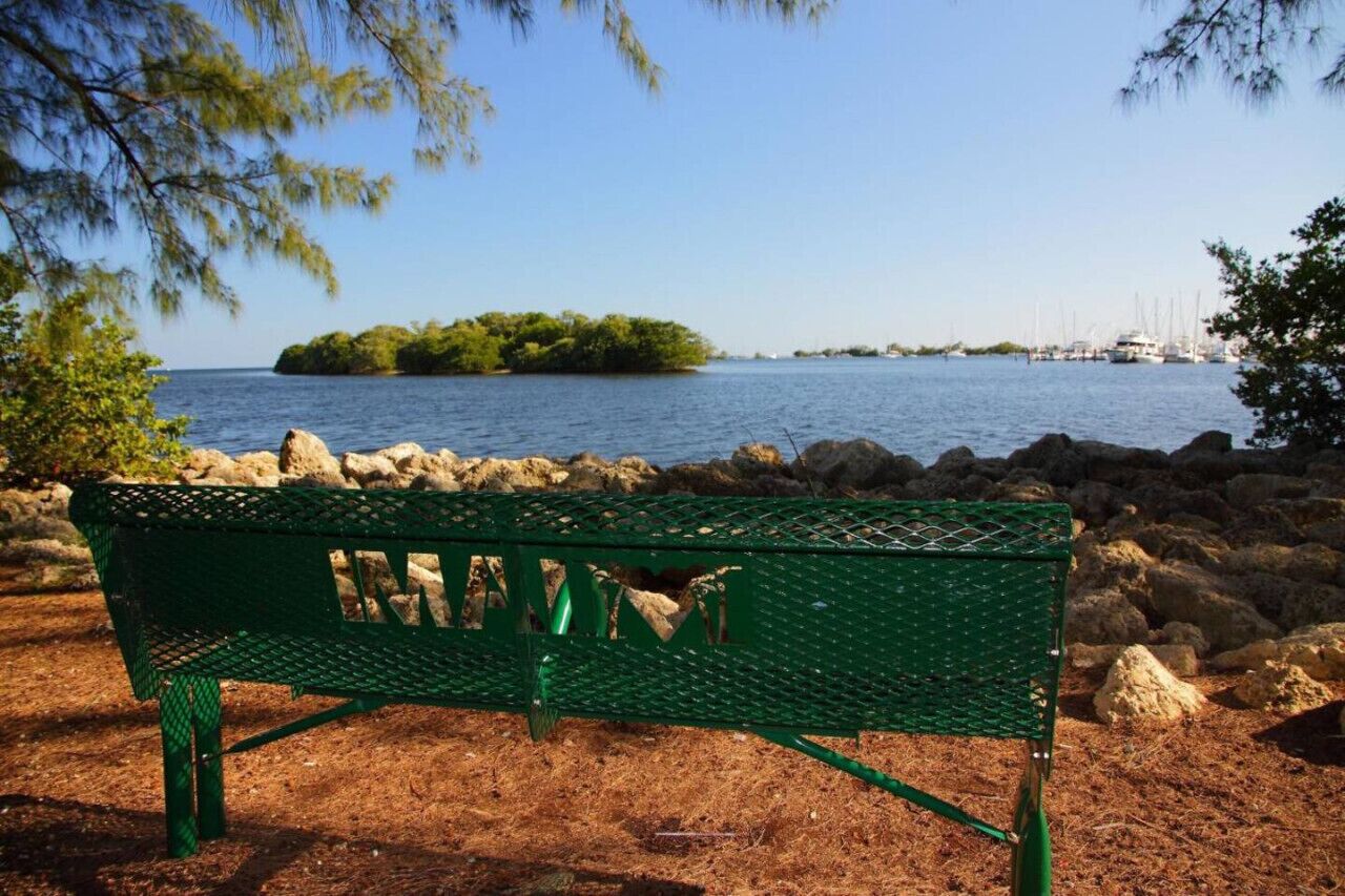 Un banco de metal verde junto a una costa rocosa, con vista a una pequeña isla y veleros a lo lejos.