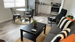 Een moderne woonkamer met een bank, salontafel en stoelen. In de achtergrond is een keuken met apparatuur en kasten te zien.