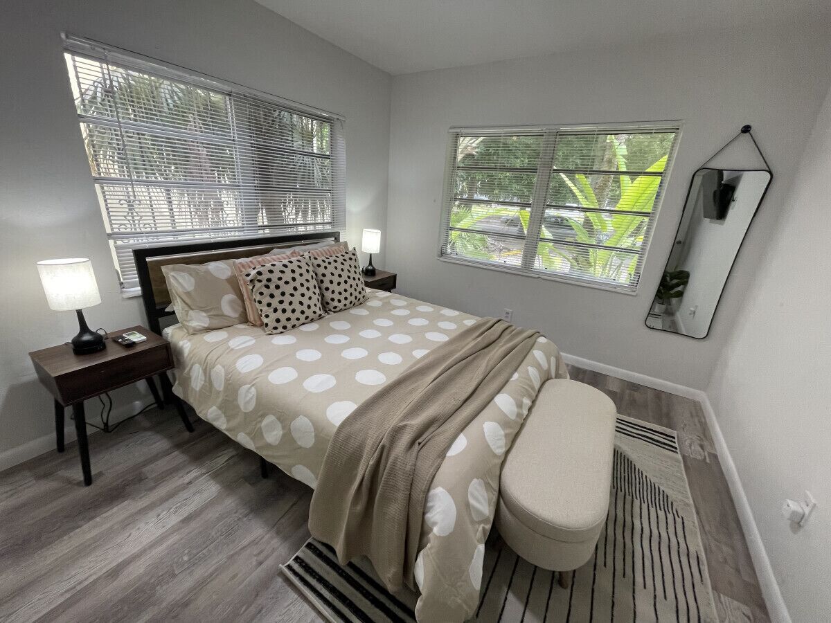 A bedroom with a bed, bedside tables, a bench, a window with blinds, and a wall-mounted mirror.