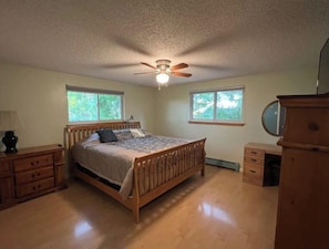 Quarto com cama de madeira, duas janelas, ventoinha no teto, cômoda e espelho.