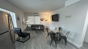 Una sala moderna con un sofá, una mesa de centro y una mesa de comedor con sillas. Hay un televisor de pantalla plana montado en la pared y un refrigerador en la esquina.
