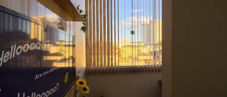 Una habitación con un ventanal grande, un televisor montado en la pared, una mesita con una planta en maceta y un ventilador de pie.
