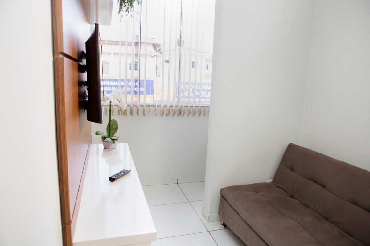 Una habitación con un sofá blanco, un televisor montado en la pared, una mesita con una planta y una ventana con persianas.