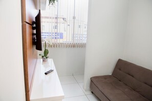 Et værelse med en hvid sofa, et fjernsyn ophængt på væggen, et lille bord med en plante og et vindue med persienner.
