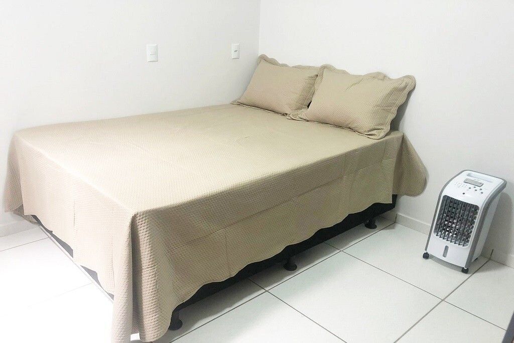 Una cama individual con colcha y almohadas color beige, un calentador junto a la cama y paredes blancas.
