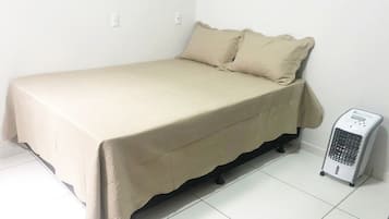 Una cama individual con colcha y almohadas color beige, un calentador junto a la cama y paredes blancas.