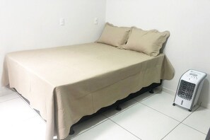En enkeltseng med et beige sengetæppe og puder, en varmeblæser ved sengegavlen, og hvide vægge.