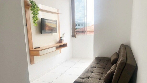 A modern living room with a sofa, a mounted TV, and a window with a view of buildings. - CONDOMÍNIO SONHARE - 202 (Anapolis)