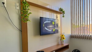 A modern living room with a mounted TV, wooden TV stand, a remote control, a small potted plant, and a standing fan. - CONDOMÍNIO SONHARE - 202 (Anapolis)