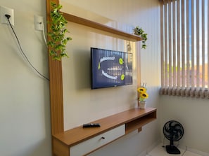 Una sala de estar moderna con un televisor montado, un mueble bajo de madera para el televisor, un control remoto, una pequeña planta en maceta y un ventilador de pie.