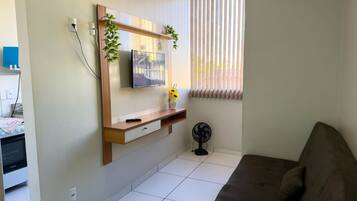 Uma sala de estar com televisão de tela plana, um móvel de madeira para TV, um vaso de planta, um ventilador de chão e um sofá.