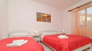 A hotel room with two beds, each with a red bedspread and white pillows, a nightstand with a lamp, and a painting of a beach scene on the wall.