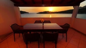 Una mesa de comedor con cuatro sillas y un jarrón de flores, en un balcón con vistas a la puesta de sol sobre el agua.