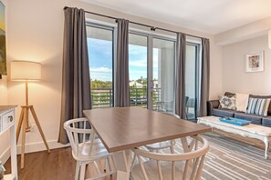 A modern living room with a wooden table, white chairs, a sofa with cushions, a floor lamp, and a view through large windows. - Charming Waterfront 1/1.5 Beautiful Sunsets at the Strand! (Sarasota)