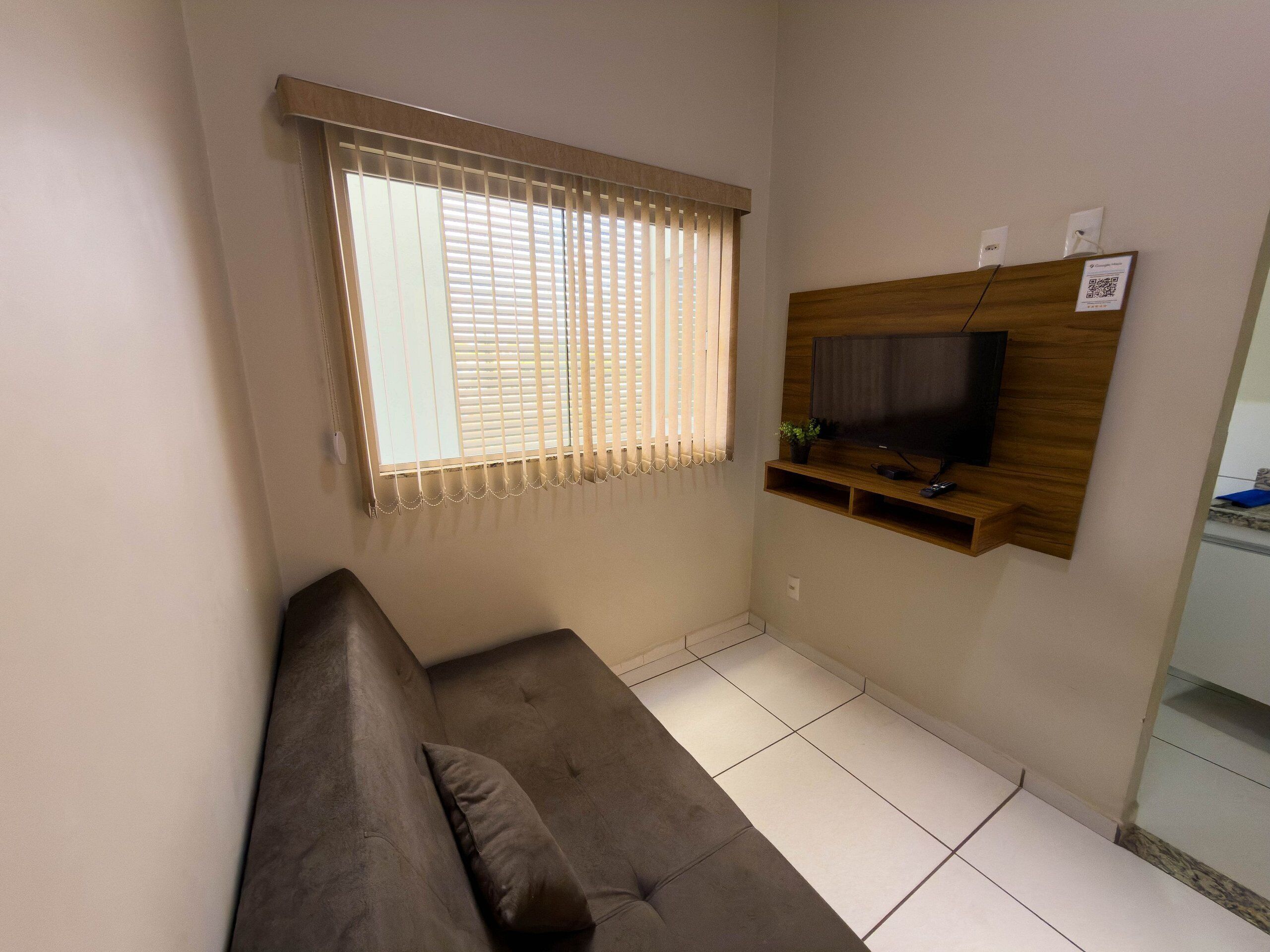 A living room with a sofa, a television mounted on a wooden wall, and a window with blinds.