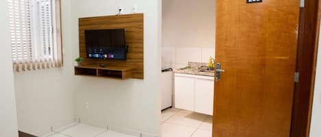 Een kamer met een aan de muur bevestigde televisie, een deur met de tekst 203, en een keukenruimte zichtbaar door een open deur.
