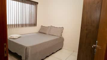 A small, neatly made bed with a plain bedspread and pillows, a window with vertical blinds, and a wooden door.