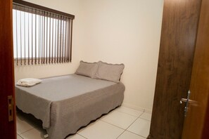 A small, neatly made bed with a plain bedspread and pillows, a window with vertical blinds, and a wooden door. - CONDOMÍNIO SONHARE - 203 (Anapolis)