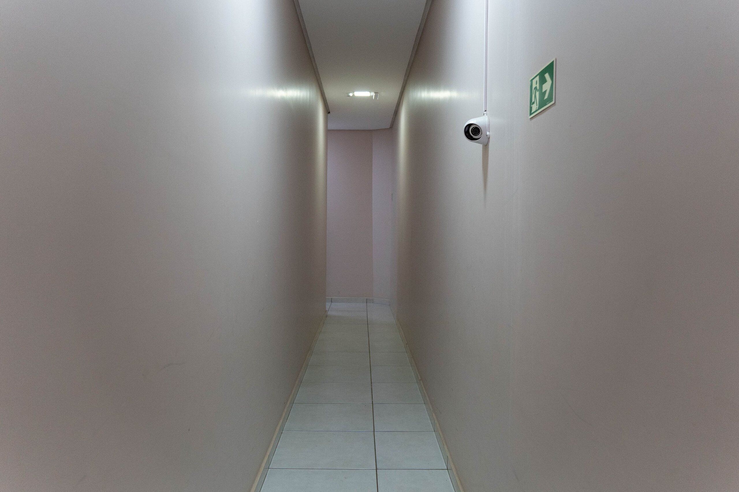 A narrow hallway with a tiled floor, a security camera, and an emergency exit sign on the wall.