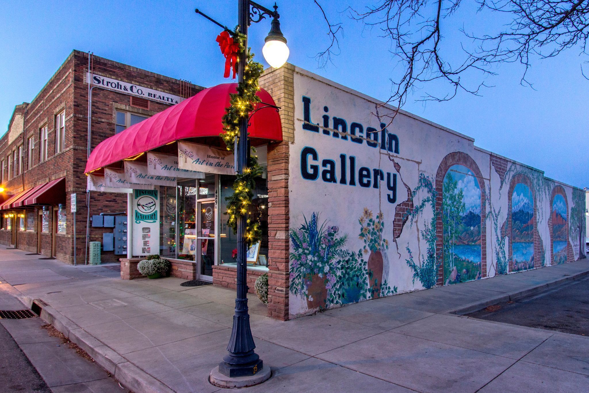 A mural of a landscape on a building's exterior wall, a street lamp with lights, and a storefront with a red awning.