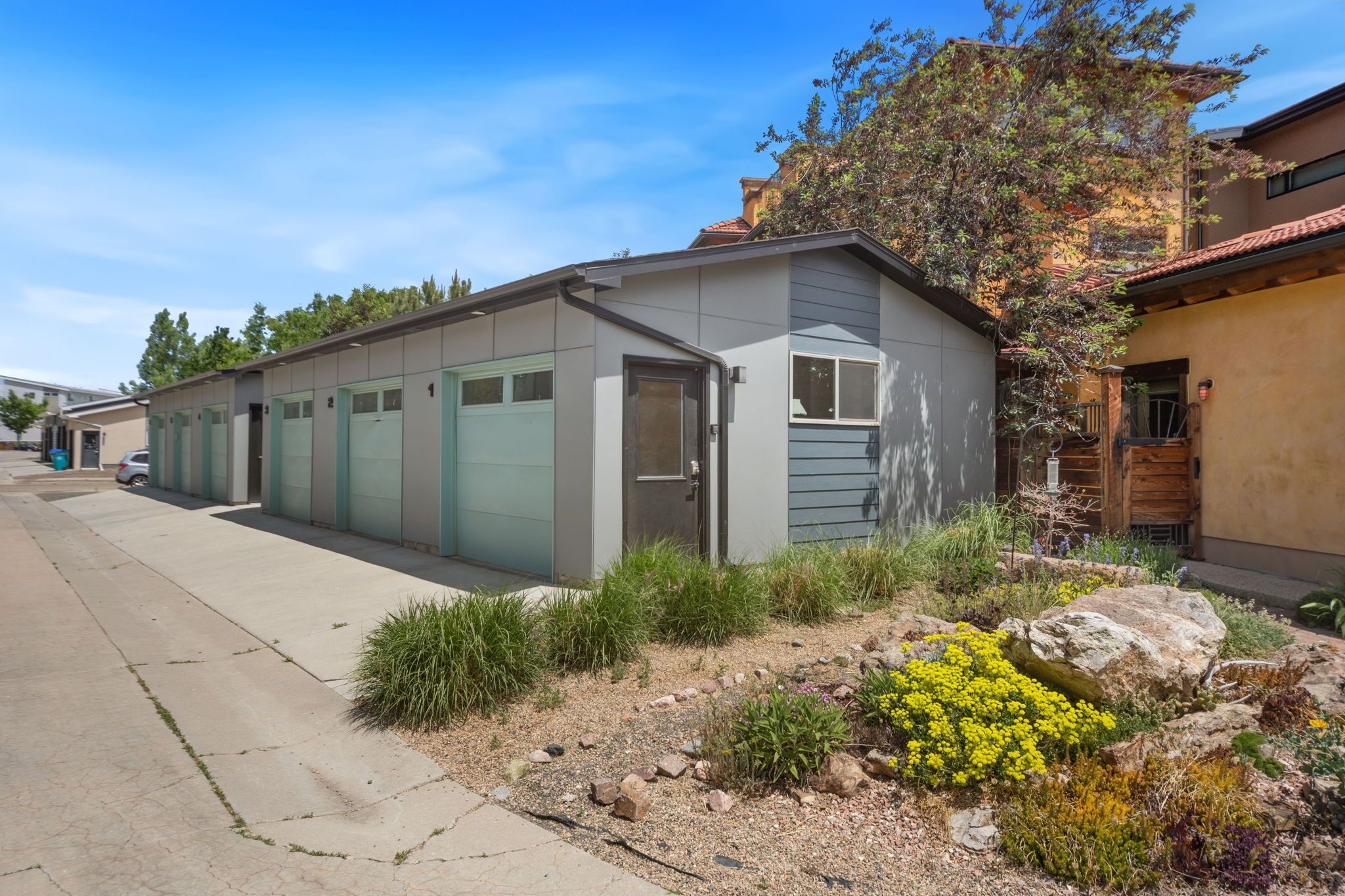 A modern building with a garage door, a small garden with rocks and yellow flowers, and a clear blue sky.