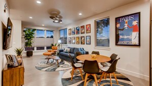 Un salon moderne comprenant un canapé, une table basse et des fauteuils, un coin repas avec une table et des chaises, et une grande fenêtre équipée de stores.