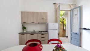 A compact kitchen with wooden cabinets, a white refrigerator, and a dining area with a red chair and a table. - 3137 Villetta Velia by Barbarhouse (Porto Cesareo)