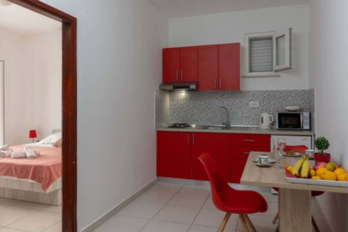 A compact kitchen with red cabinets, a microwave, and a sink. There's a dining table with a red chair and a fruit bowl.