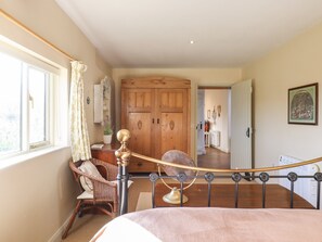 A bedroom with a large wooden wardrobe, a wicker chair, a bed with a pink blanket, and a view of a hallway leading to a kitchen.