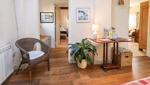 Una habitación acogedora con una silla de mimbre, una mesa de madera con una lámpara, una planta en maceta y una cama al fondo.