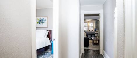 Un pasillo que conduce a un dormitorio con cama y mesita de noche, una zona de comedor con mesa y sillas, y una cocina visible al fondo.