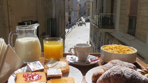 A breakfast table with a pitcher of milk, a glass of orange juice, a cup, a bowl of cereal, pastries, and a view of a street with buildings and people. - Mammasisi (Lecce)