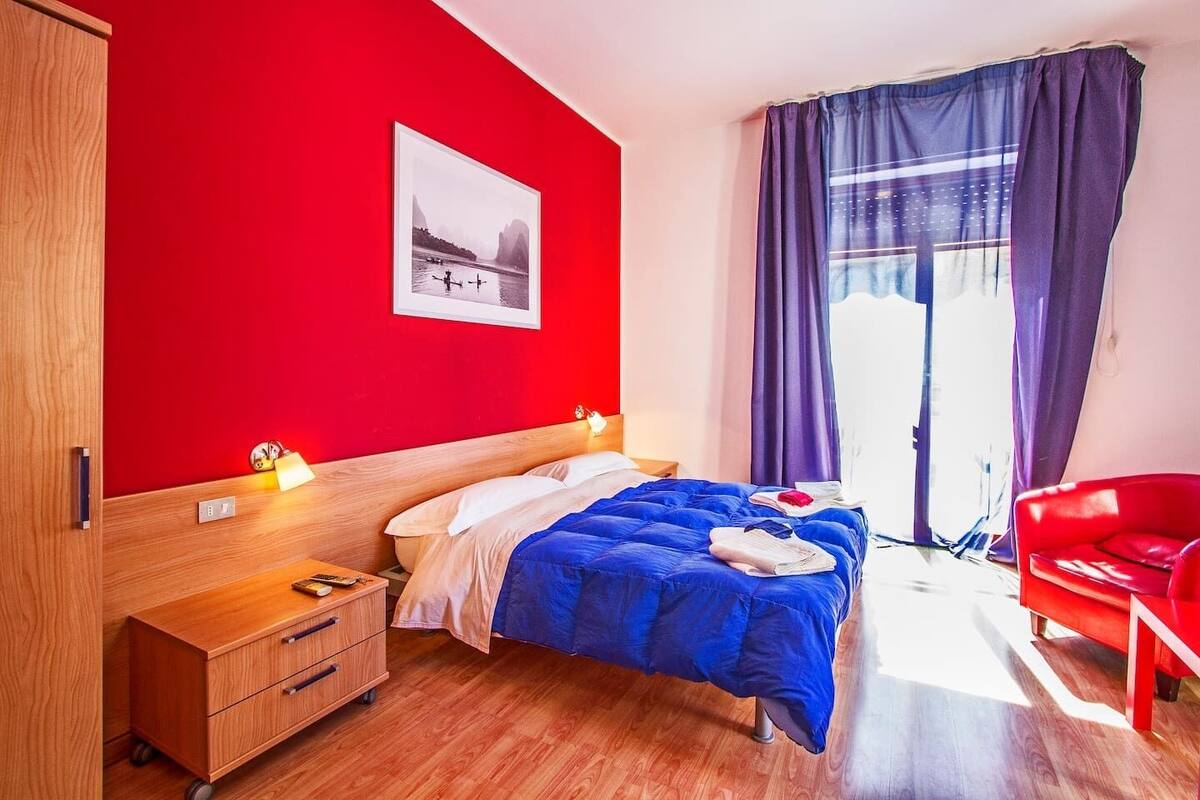 Una habitación de hotel moderna con un armario de madera, una cama con ropa de cama azul, una silla roja y un ventanal con cortinas moradas.