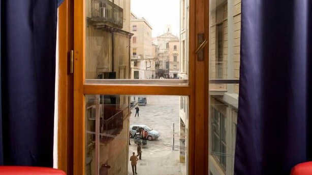 A room with a large window, a red chair, a table with a tray of breakfast items, and a view of a street with buildings and people. - Mammasisi (Lecce)