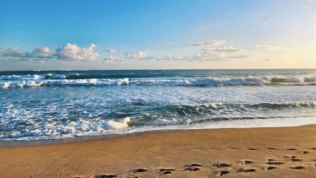 Ein Strand mit Wellen, blauem Himmel und Fußabdrücken im Sand.