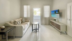 Ein modernes Wohnzimmer mit einer Couch, einem kleinen Tisch, einem Fernseher auf einem Schrank und einem Fenster mit Vorhängen.