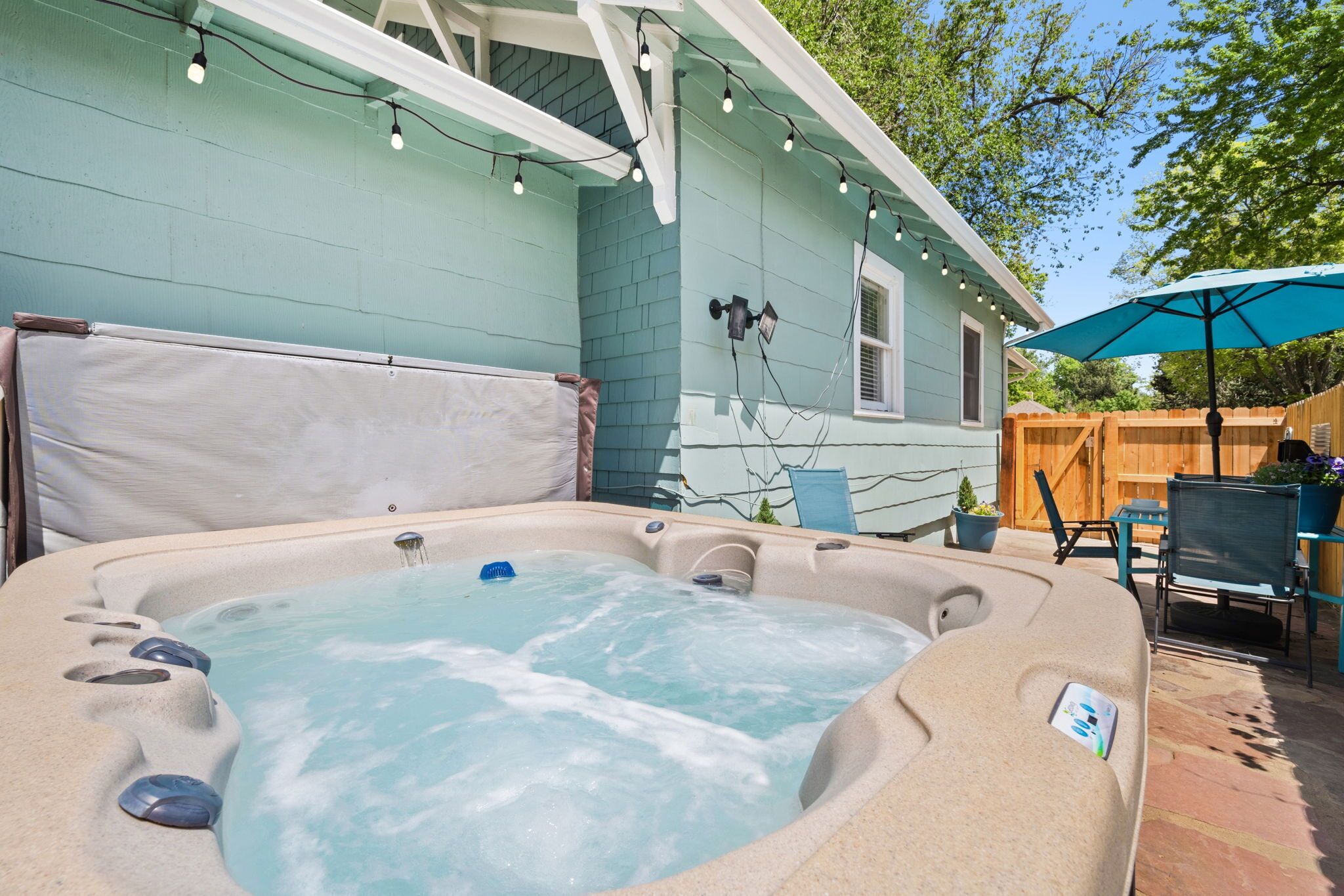 A hot tub in a backyard with a wooden fence, string lights, and a blue umbrella.