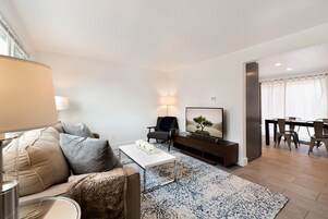 A modern living room with a sofa, coffee table, and TV. There is a dining area with a table and chairs.