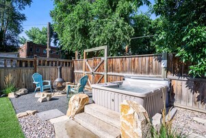 Un patio trasero con un jacuzzi, dos sillas Adirondack azules, una valla de madera y un camino de piedra.