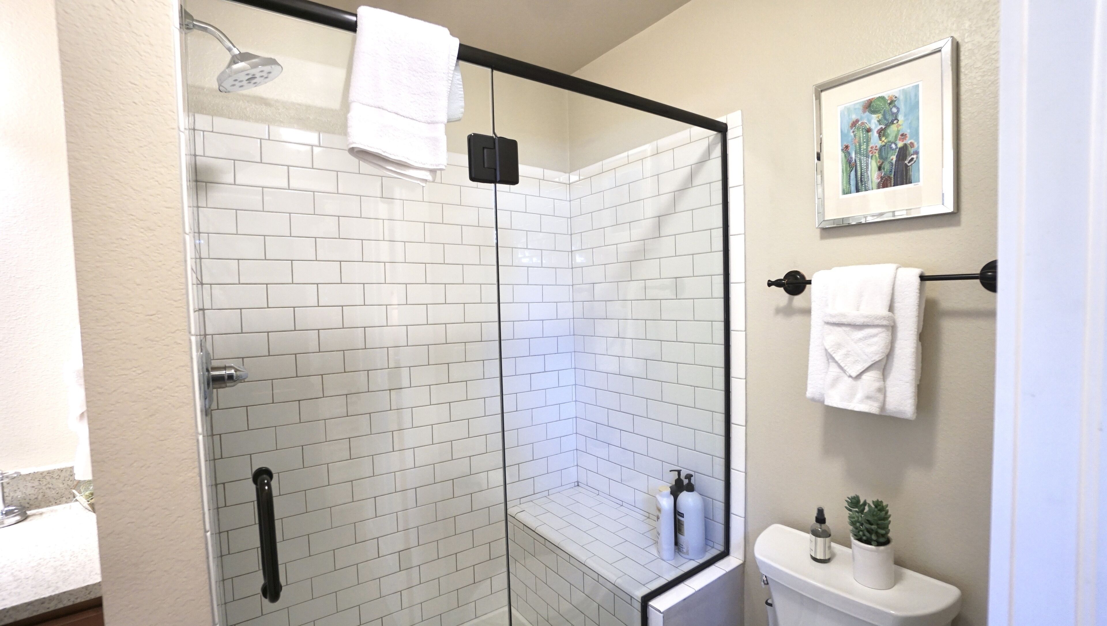 A modern bathroom with white subway tiles, a glass shower enclosure, a toilet with a potted plant, and a framed picture on the wall.