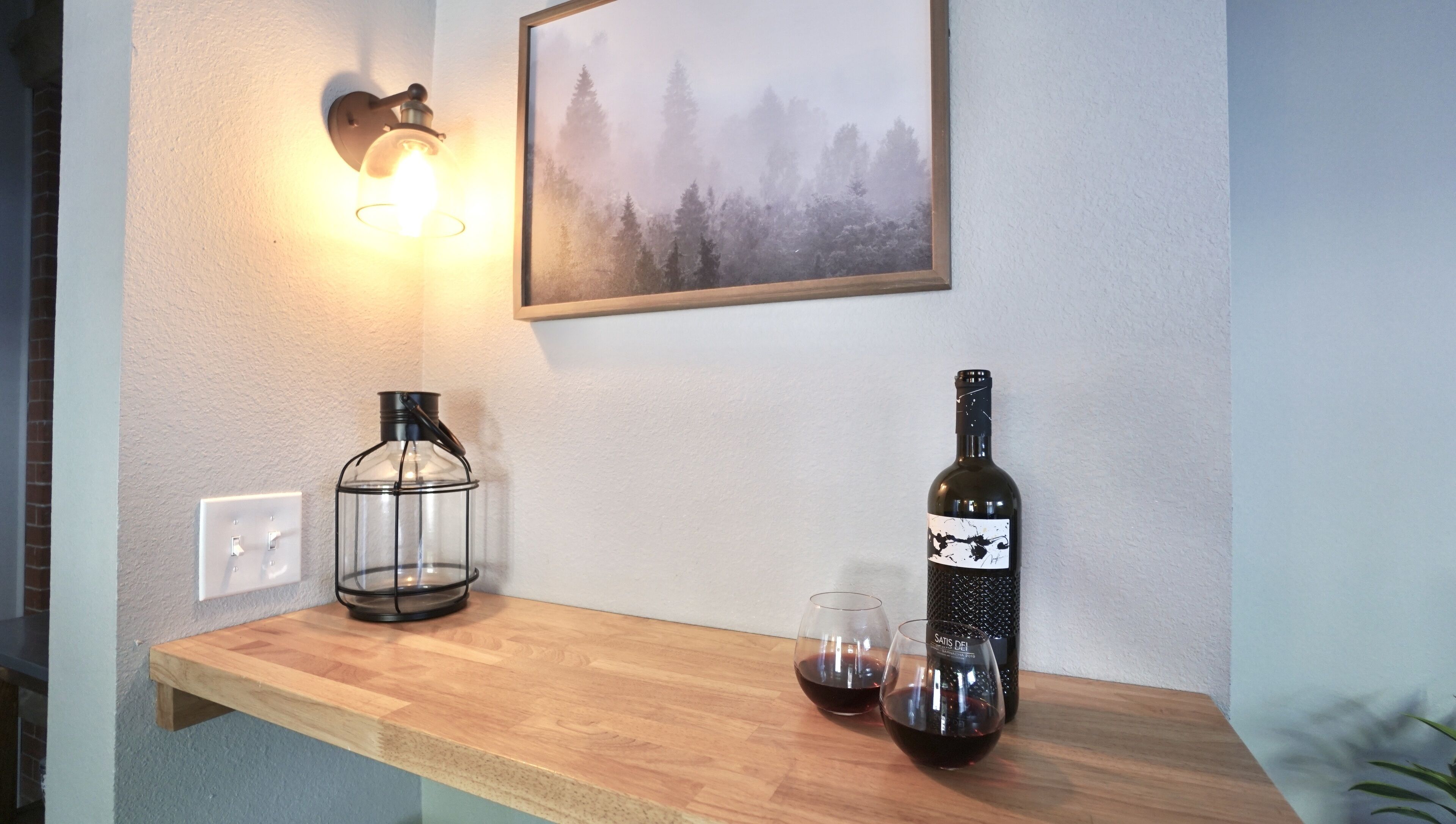 A wooden shelf with a wine bottle and two glasses, a wall-mounted light, and a framed picture.