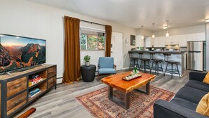 A modern living room with a TV, sofa, coffee table, and a kitchen area with bar stools and a refrigerator. - Butterscotch Suite Old Town Stay with Sunny Style (Fort Collins)