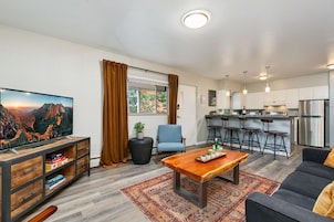 A modern living room with a TV, sofa, coffee table, and a kitchen area with bar stools and a refrigerator.